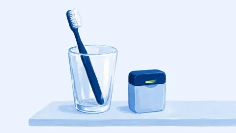 Un cepillo de dientes en un vaso y una cajita de hilo dental lisa, sobre una repisa.